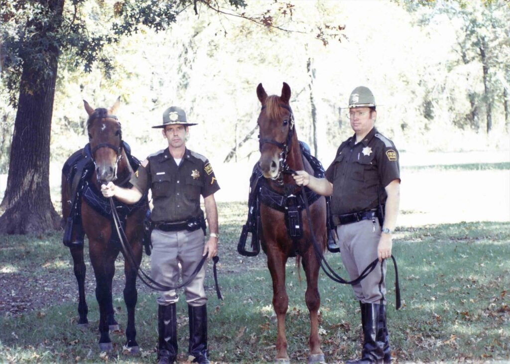 Two officers stand holding their horses under the trees
