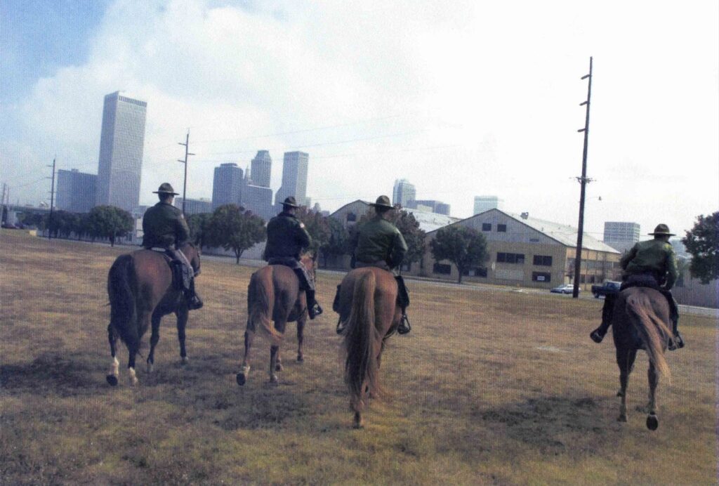 Four mounted officers ride across a field toward the Tulsa skyline